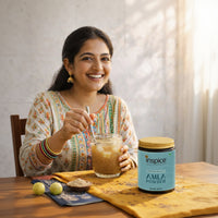 Woman holding a glass of amla powder drink with a container of Inspice Amla Powder on a table.